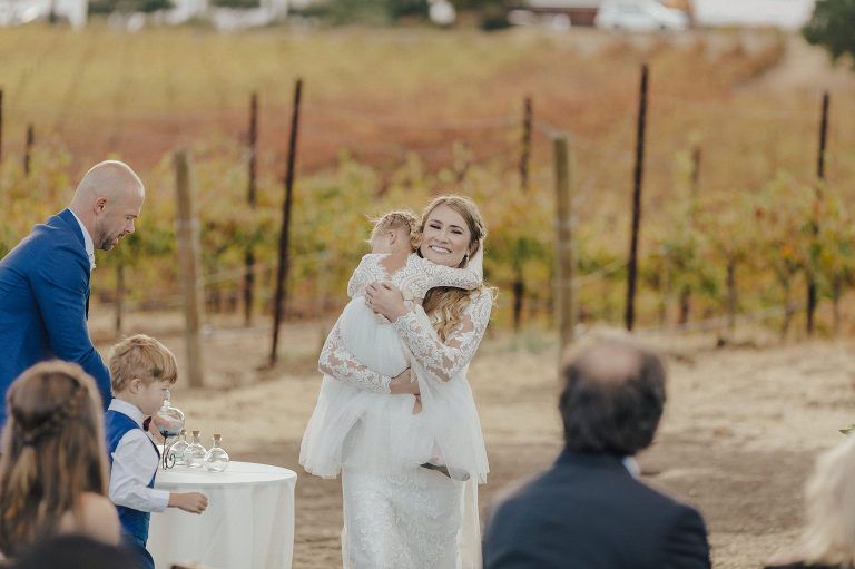 Bride embraces a child during an outdoor wedding ceremony, vineyard in the background.