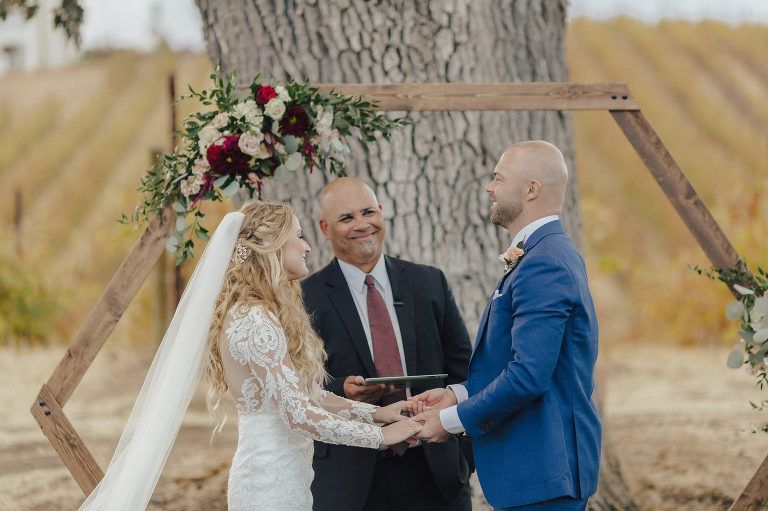 Couple exchanging vows at a wedding ceremony, under a wooden arch decorated with flowers, with an officiant present.