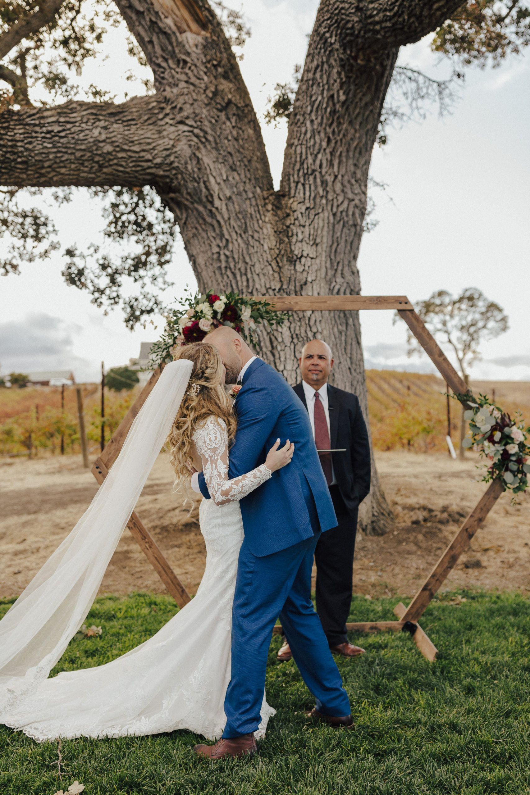 Bride and groom embrace at outdoor wedding ceremony. Wooden hexagon arch, officiant nearby, large tree in background.
