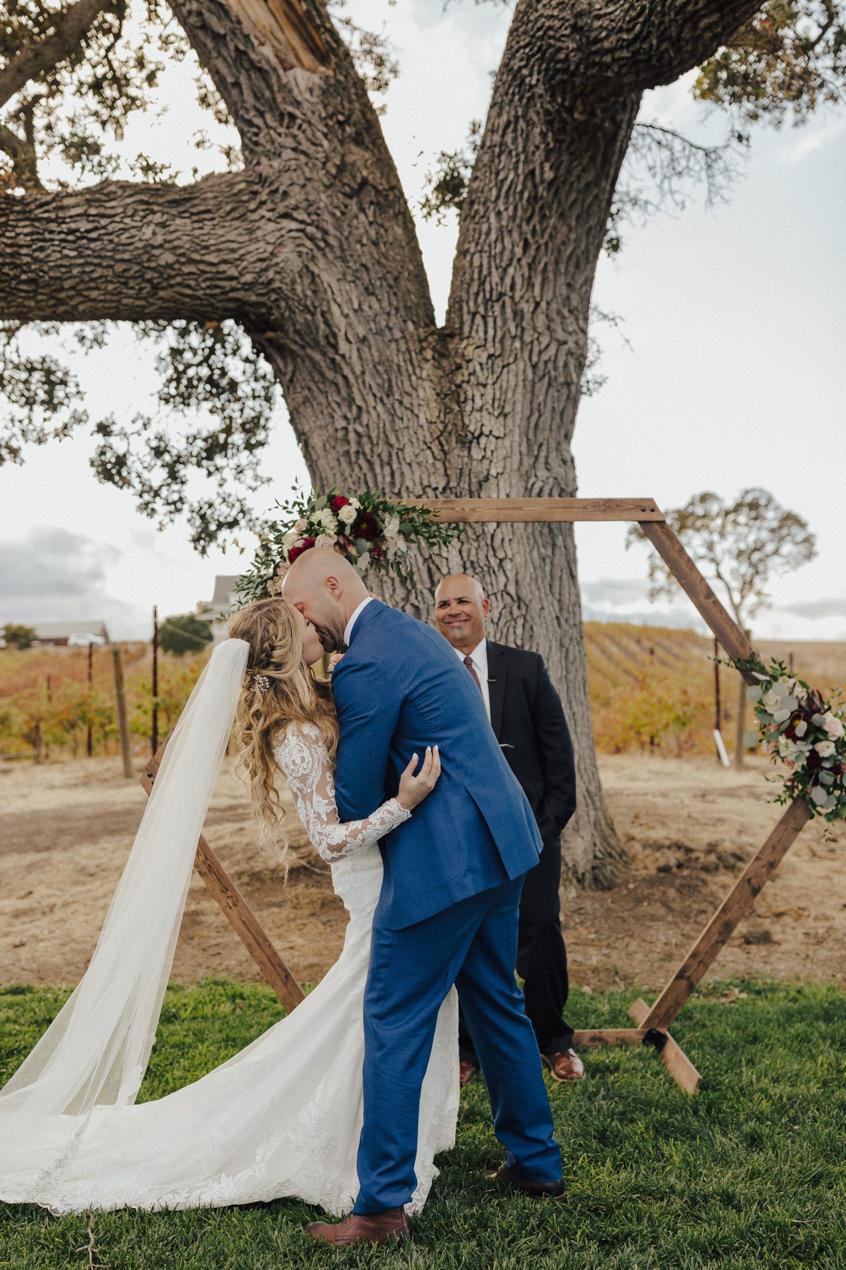Bride and groom kissing under wooden arch near large tree; man in suit watches.