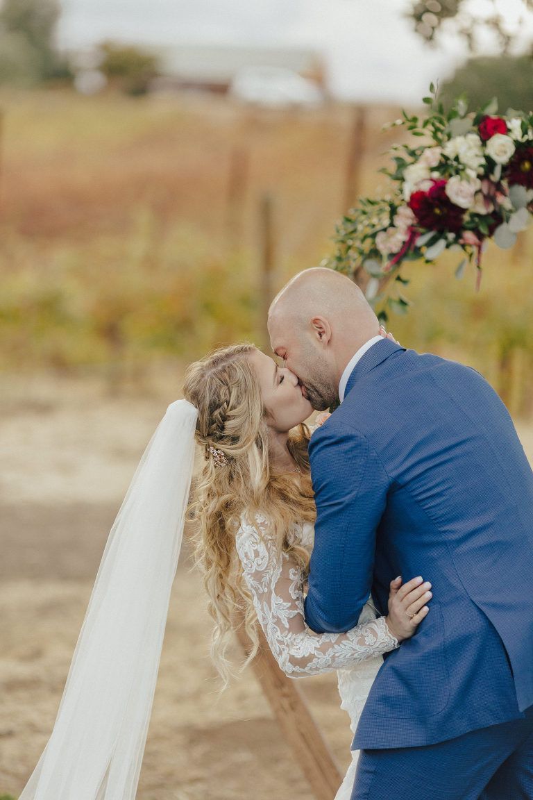 Bride and groom kissing under flower arch at outdoor wedding.