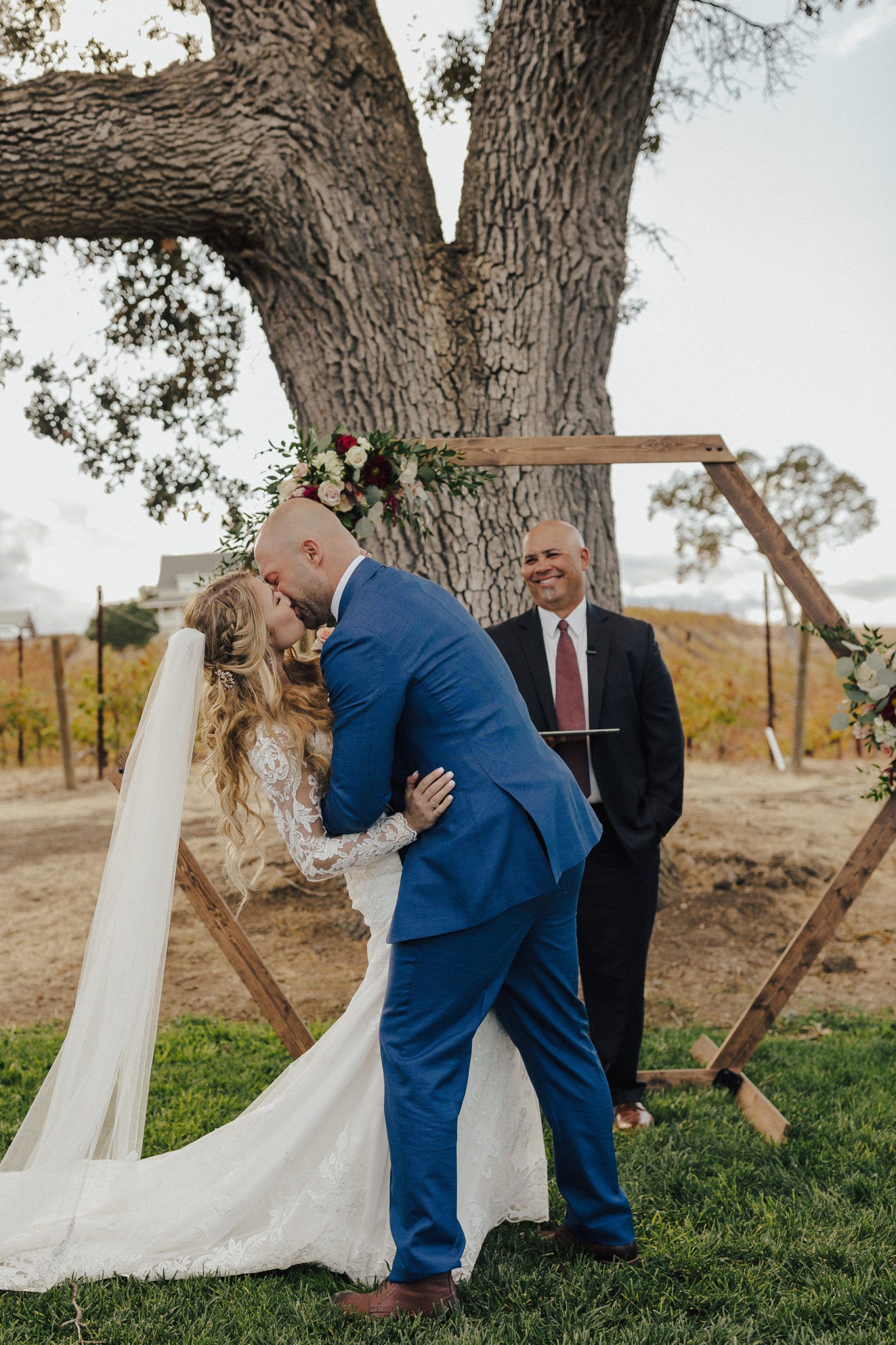 Couple kissing at wedding ceremony, wooden arch, officiant standing by tree in outdoor setting.