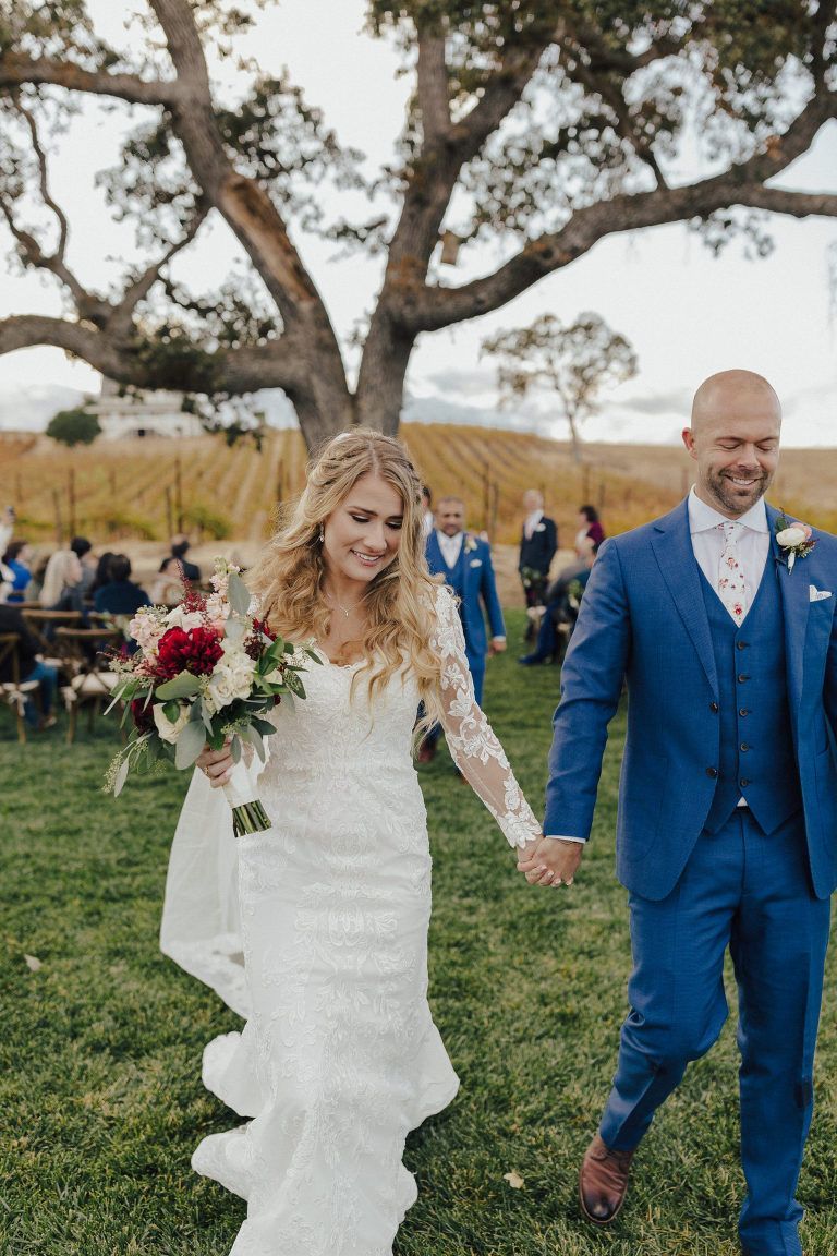 Newlyweds walking hand-in-hand after ceremony, bride in white dress, groom in blue suit, outdoor setting.