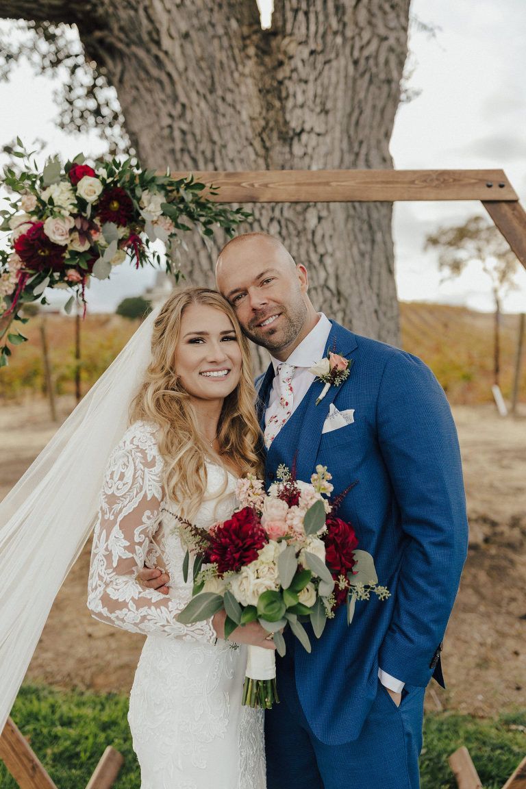 Bride and groom pose outdoors. She wears a white dress, veil. He wears a blue suit. Flowers and a wooden arch are behind them.