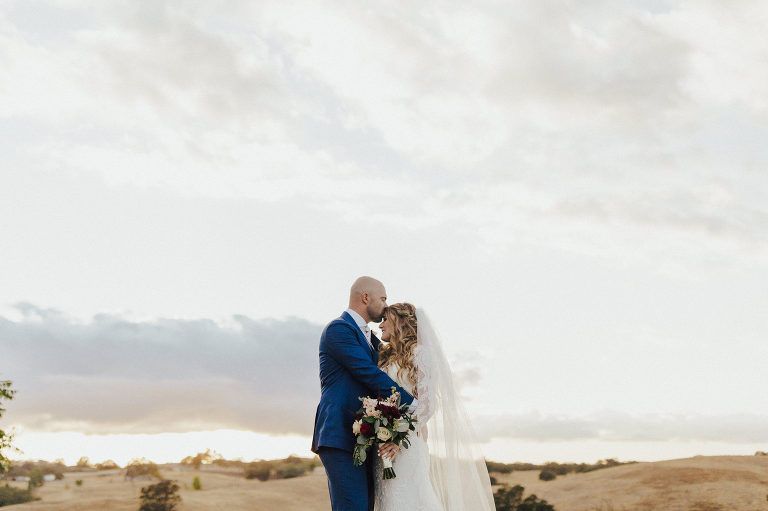 Couple embracing outdoors at sunset; man in blue suit, woman in white dress, veil.