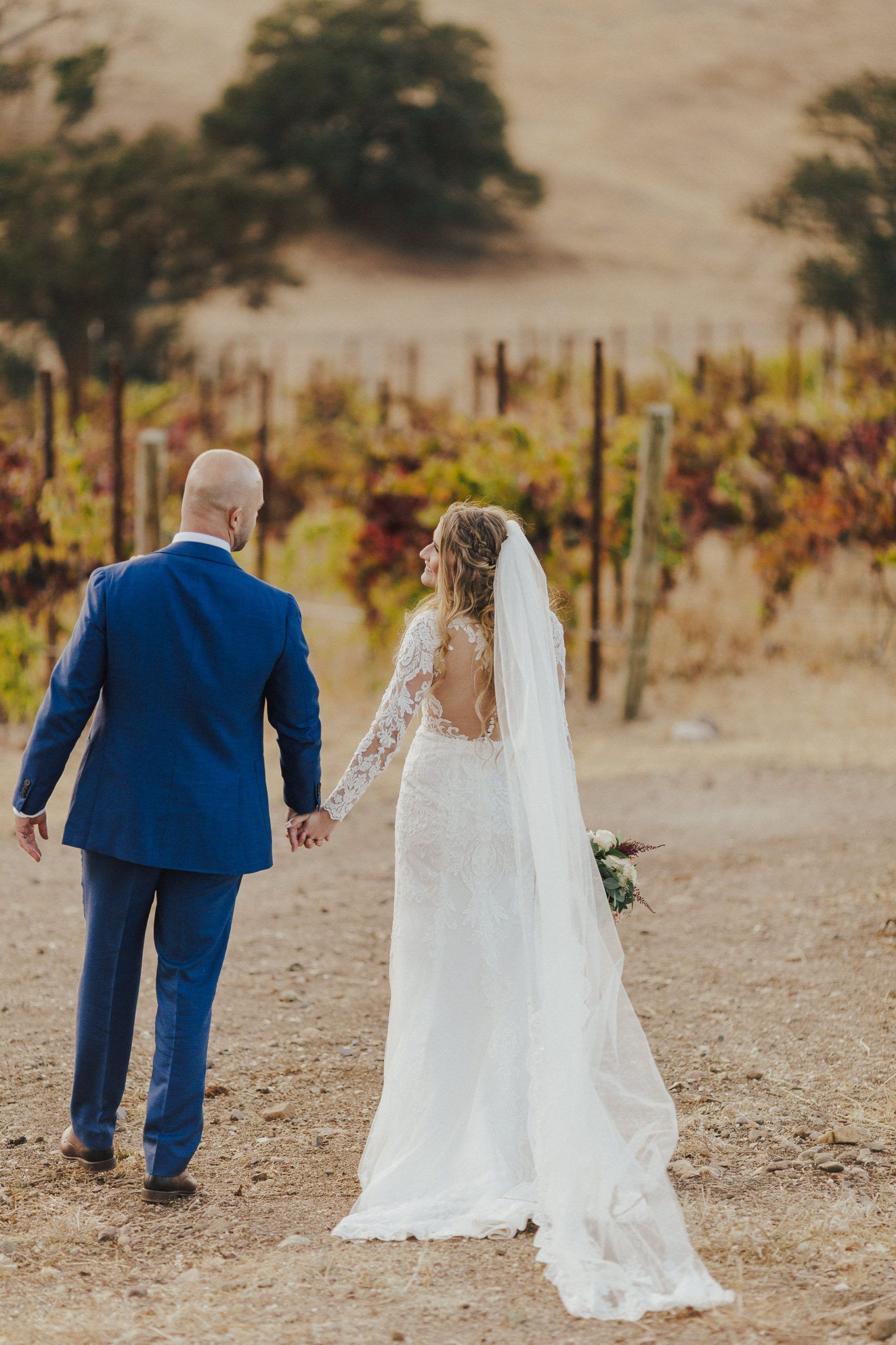 Bride and groom walking hand-in-hand in vineyard after ceremony. Bride wears a white lace gown and veil, groom a blue suit.