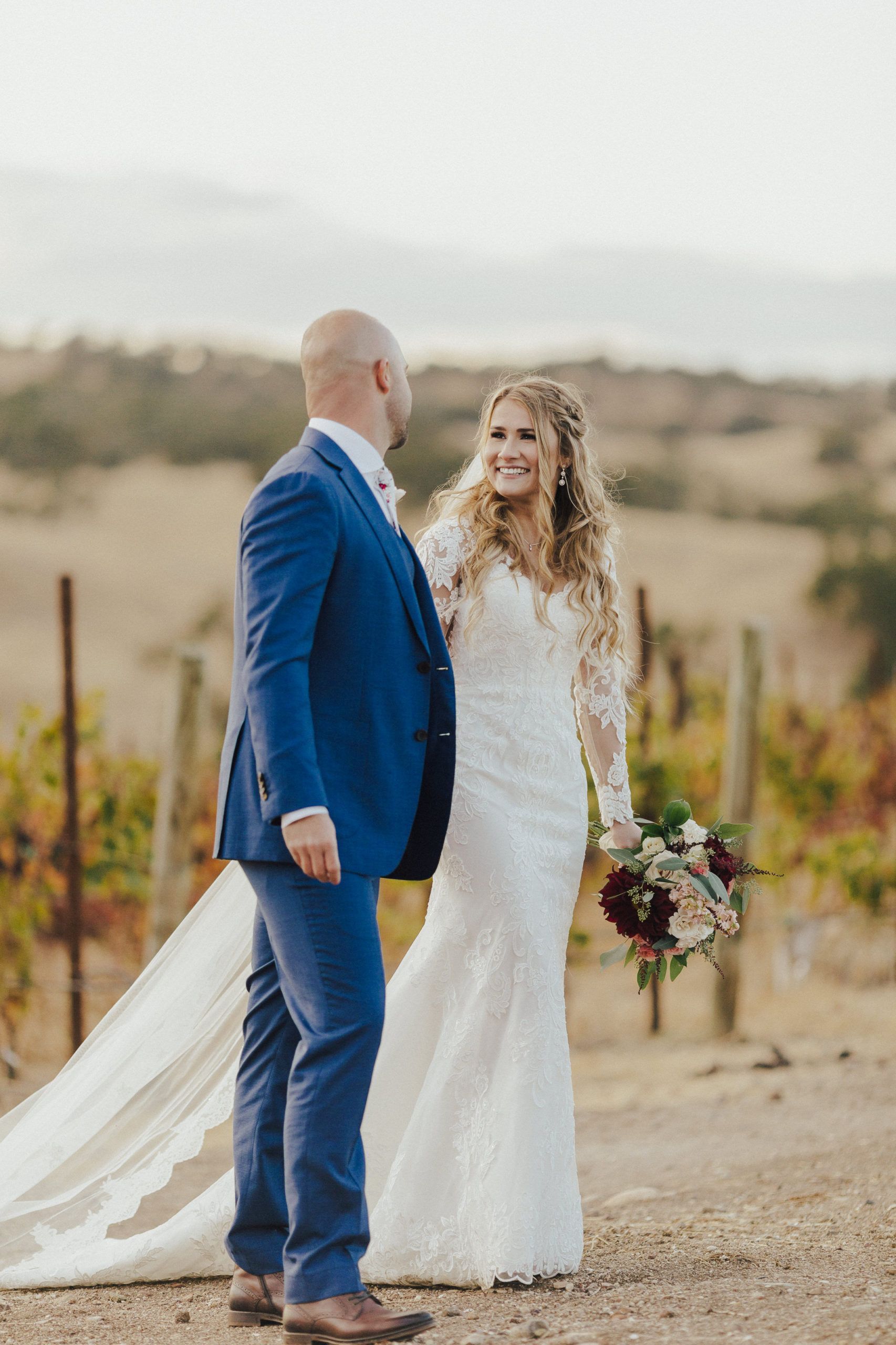 Bride and groom smiling at each other; the bride in a long white dress, groom in blue suit. Outdoor setting with vineyard.
