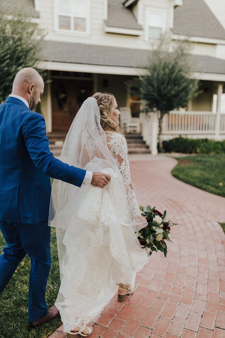 Bride and groom walk toward a house, she in a white dress, he in a blue suit, on a brick path.