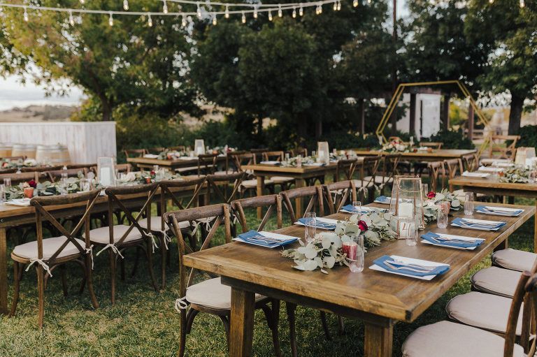 Outdoor wedding reception setup with wooden tables, chairs, and floral centerpieces; string lights overhead.