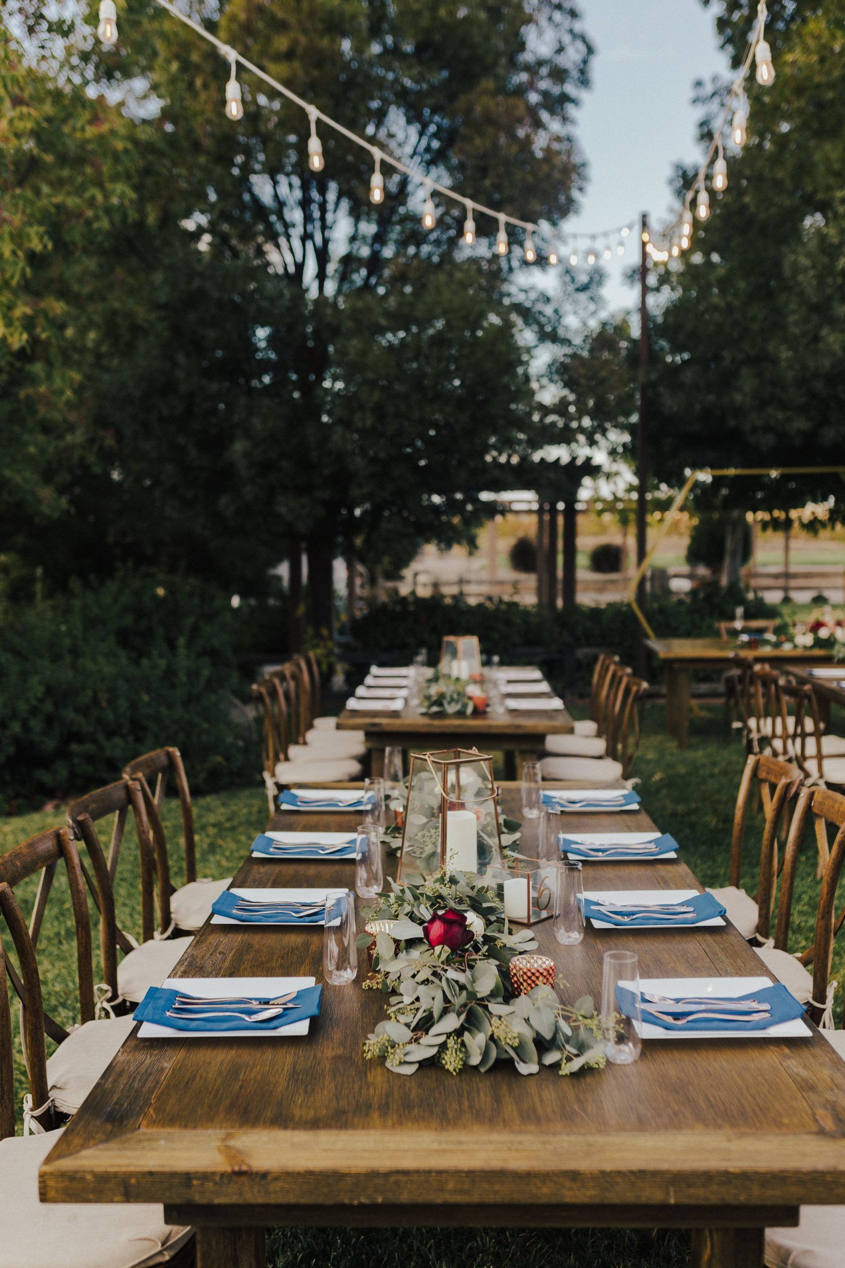 Long wooden tables set for an outdoor event, decorated with floral centerpieces and string lights.