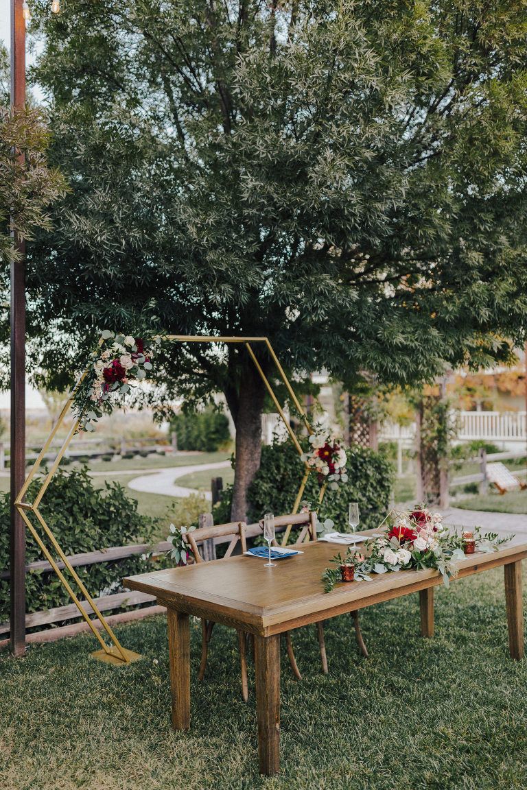 Wedding reception setup with wooden table, floral arrangements, and gold geometric backdrop outdoors.