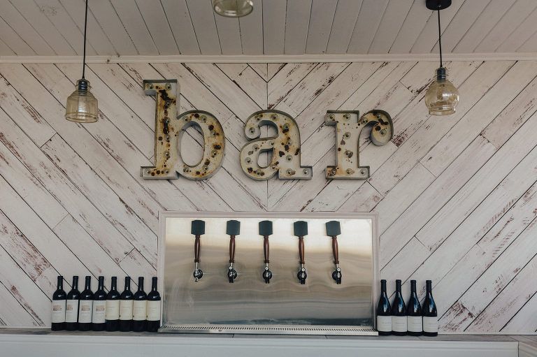 Bar sign on a whitewashed wood wall, with beer taps and bottles of wine below.