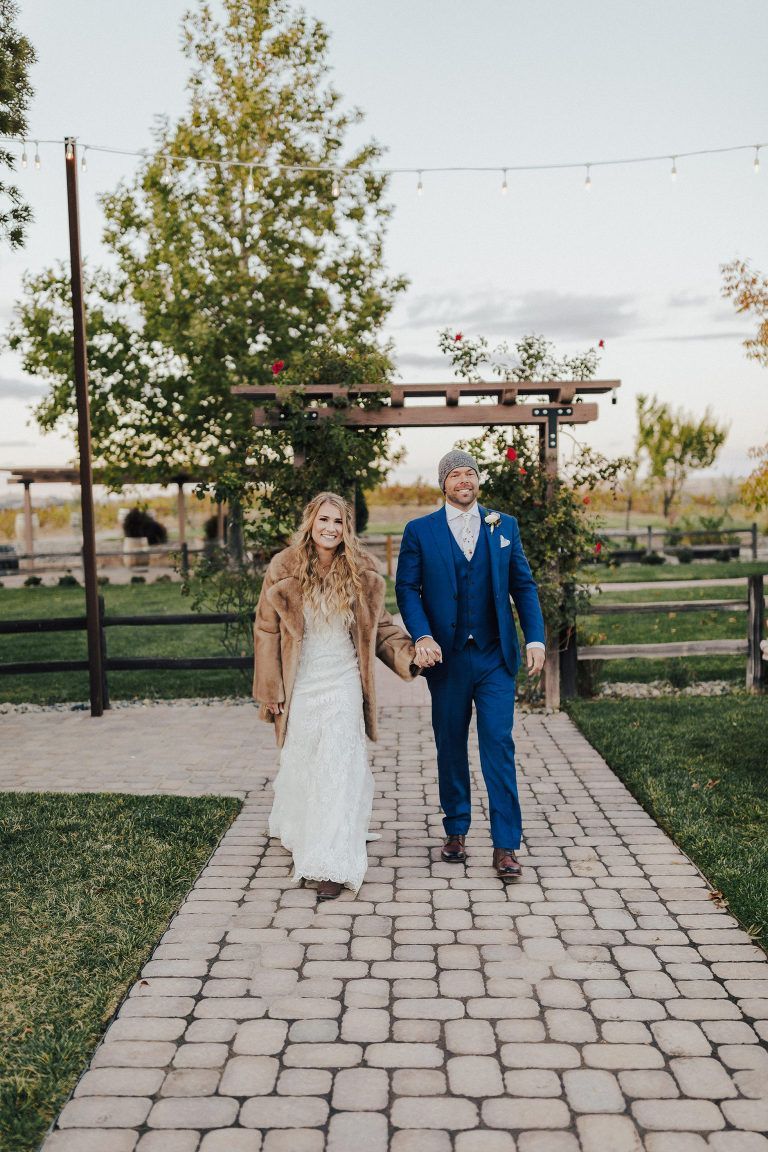 Bride and groom walking hand-in-hand down a brick path, she in a white dress and fur, he in a blue suit.