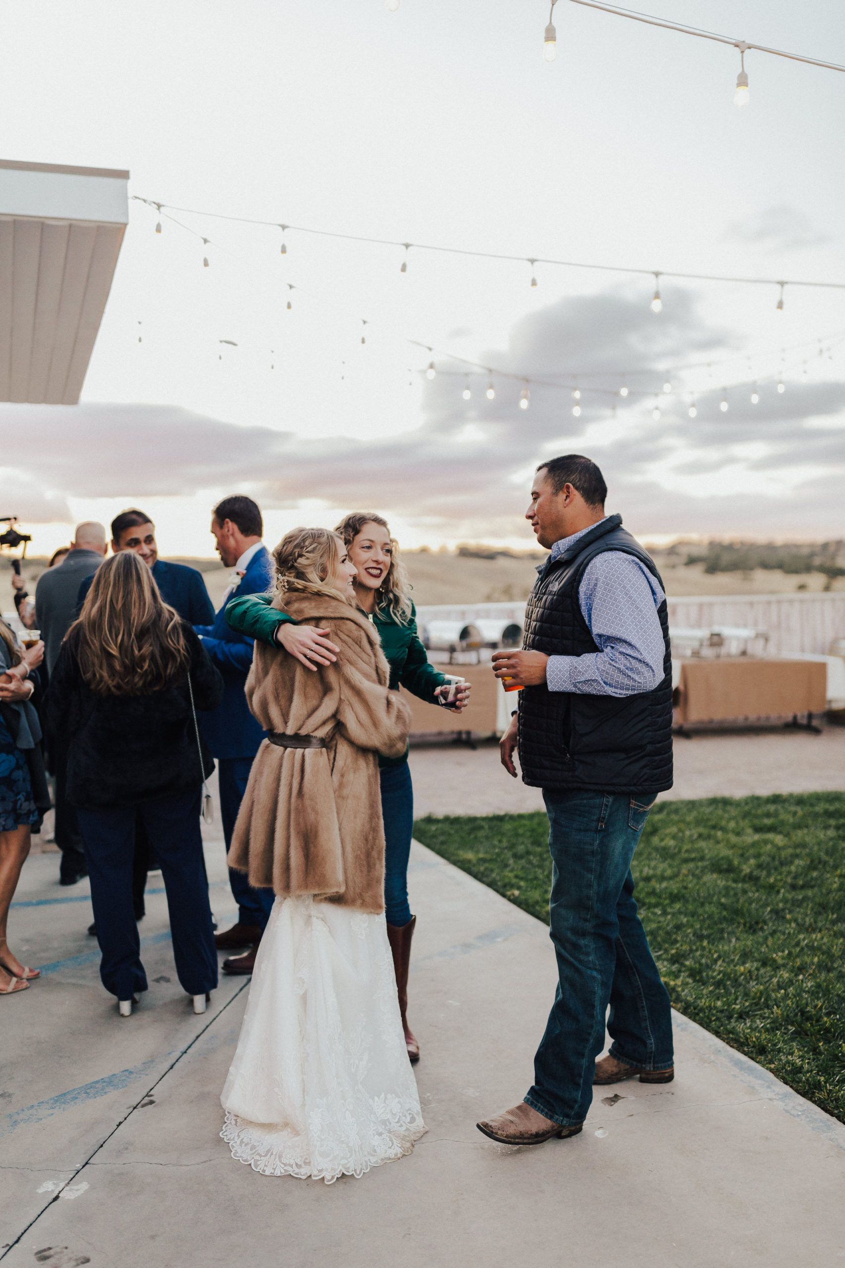 People embracing at an outdoor event with string lights. A person in a tan coat and white dress is hugged by another. A man holds a drink nearby.