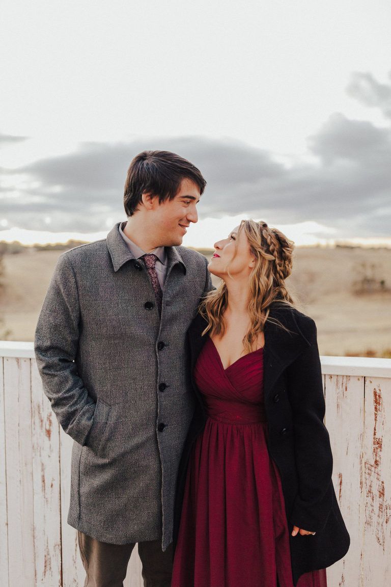 Couple gazing at each other. Man in grey coat, woman in red dress, standing by white fence. Overcast sky.
