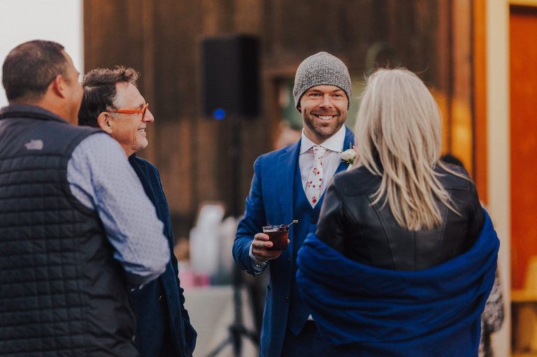 Group of people socializing outdoors; man in blue suit and hat smiles, holding a drink.