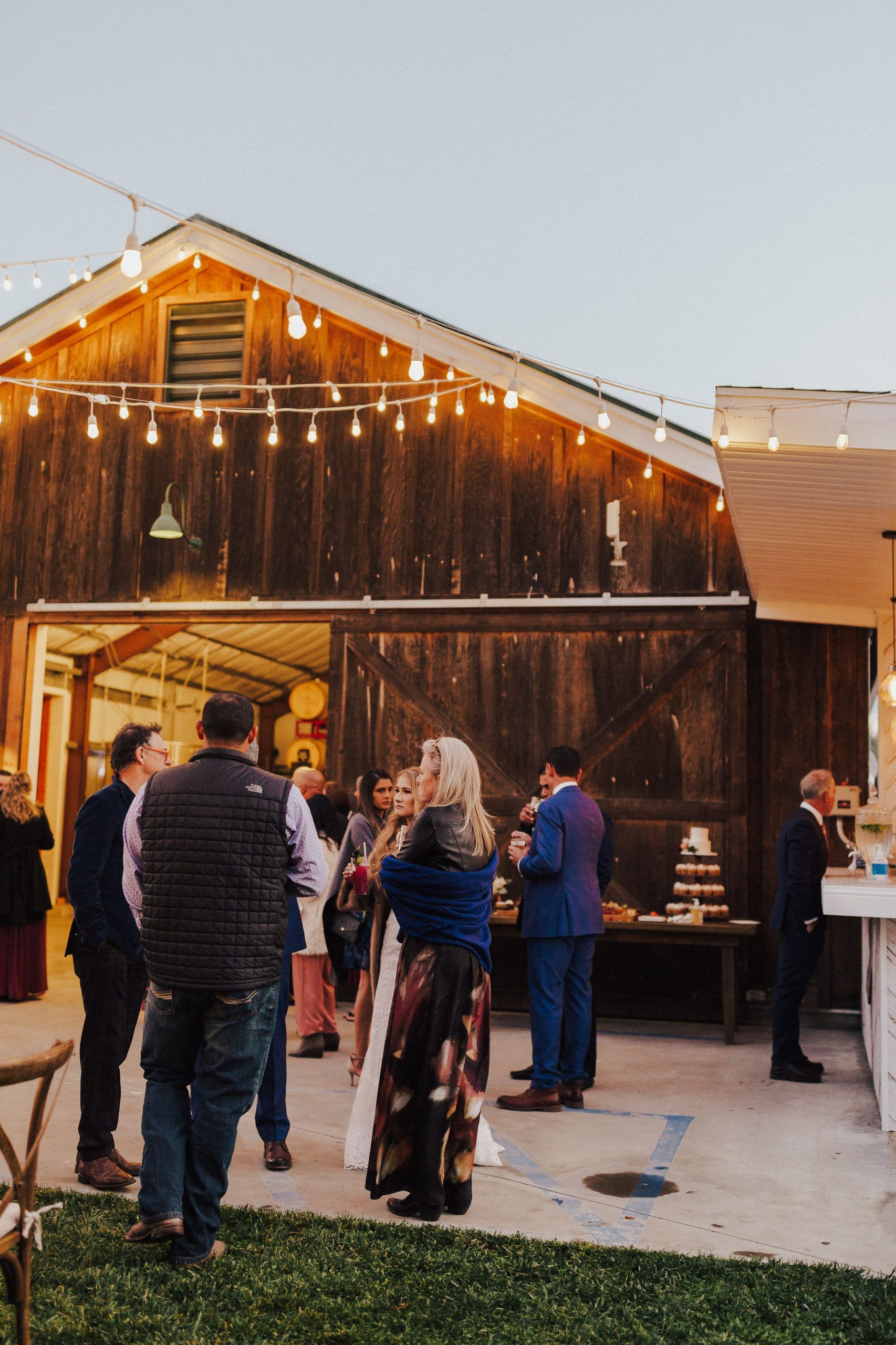 People at a wedding reception outside a wooden barn with string lights, enjoying food and drinks.