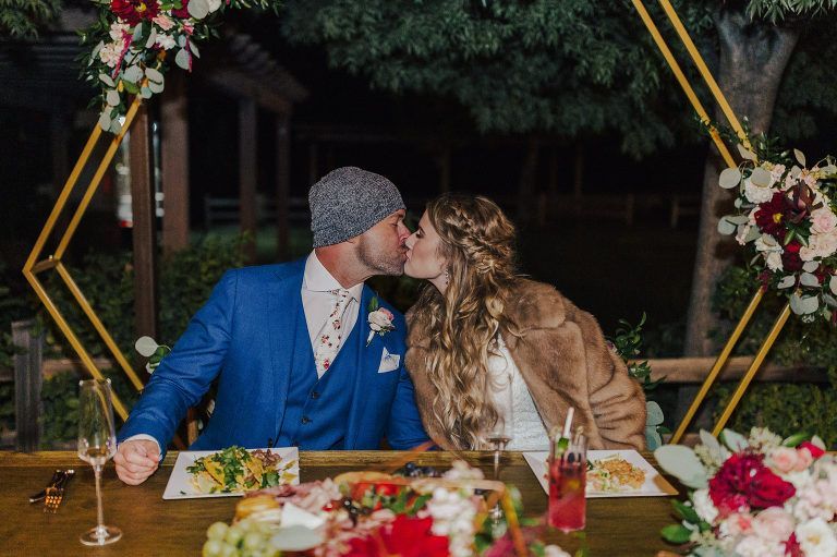 Couple kissing at a wedding reception. They sit at a decorated table, with a gold geometric arch behind them.