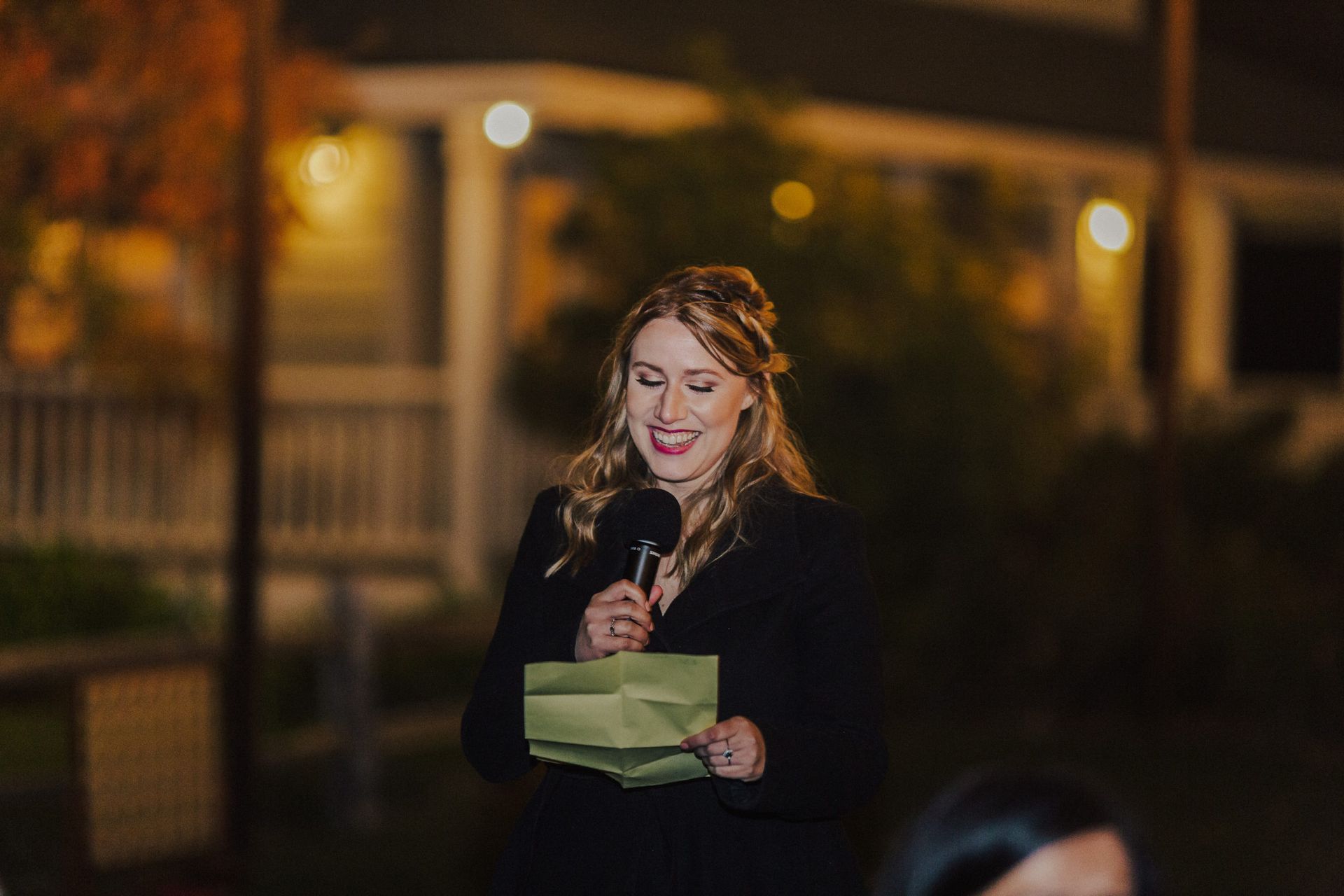 Woman speaking into microphone, holding notes outdoors at night, smiling.