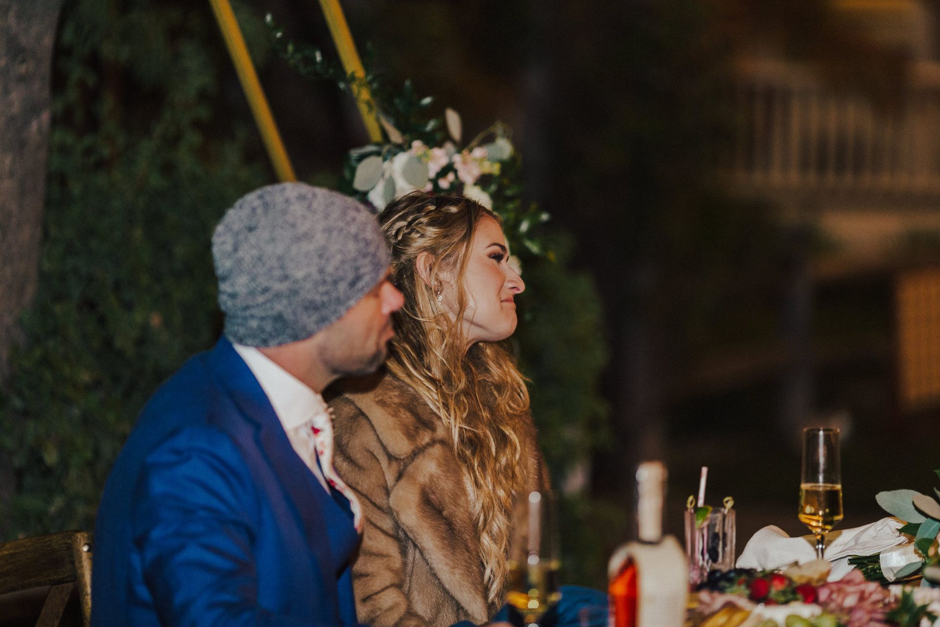 A couple at an outdoor event. Man in blue blazer and gray beanie, woman in fur coat, looking towards flowers.