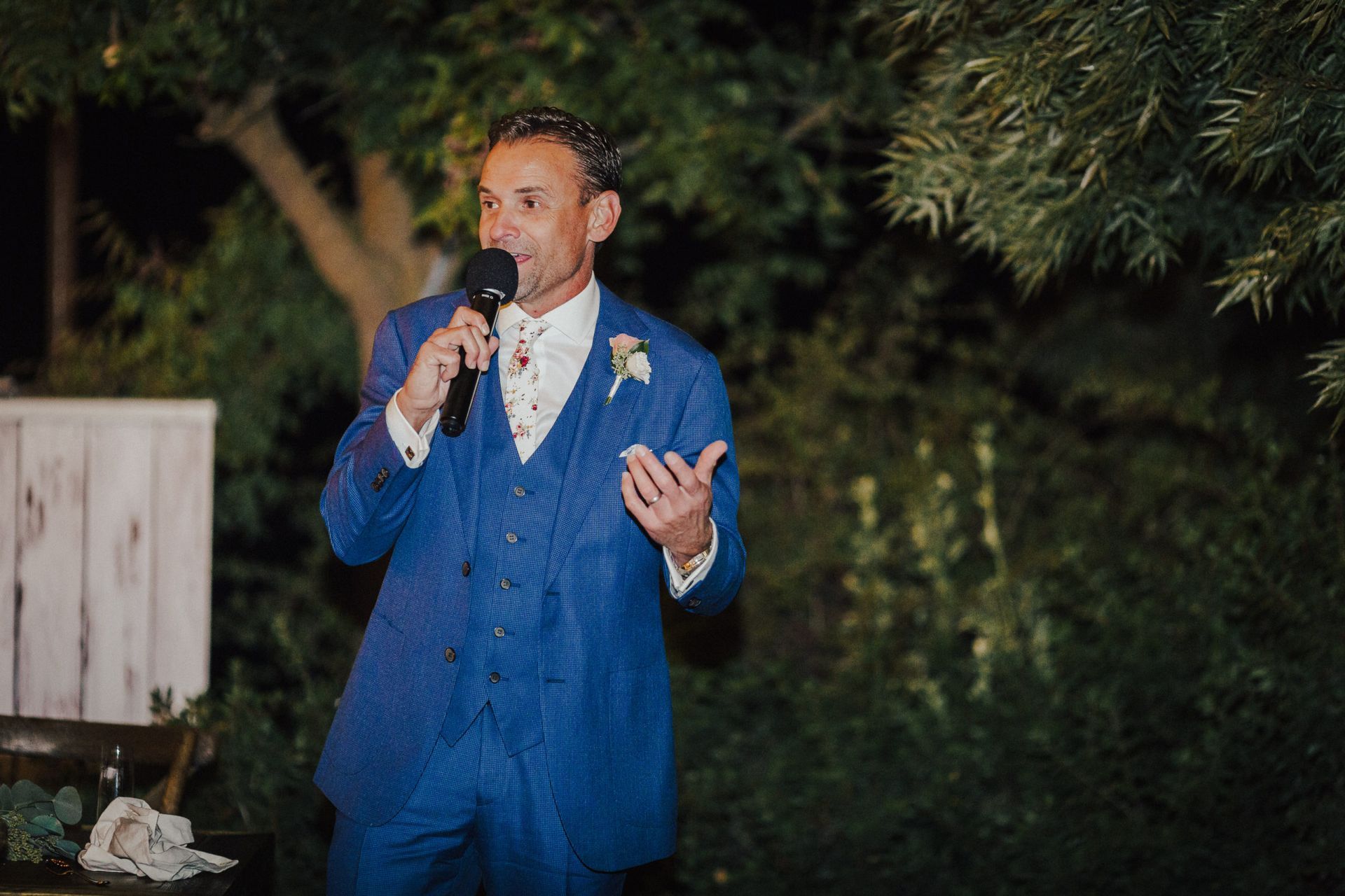 Man in blue suit speaks into microphone outdoors at night.