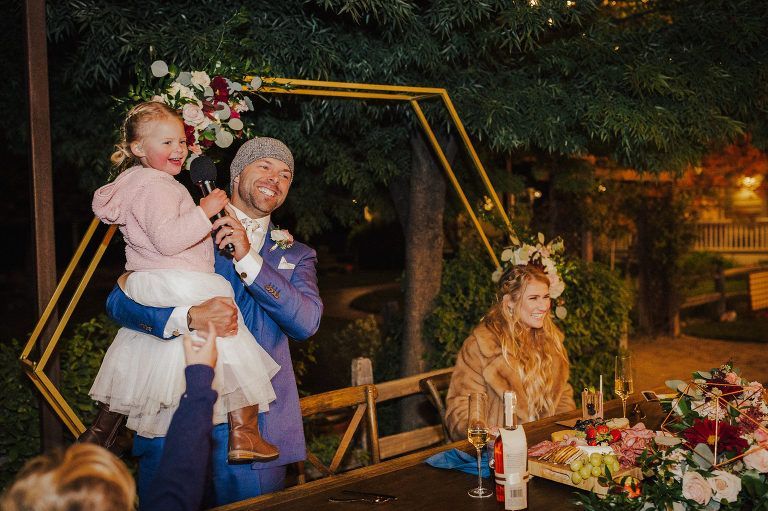 Man holding child with microphone, giving a speech at a wedding, bride seated nearby.