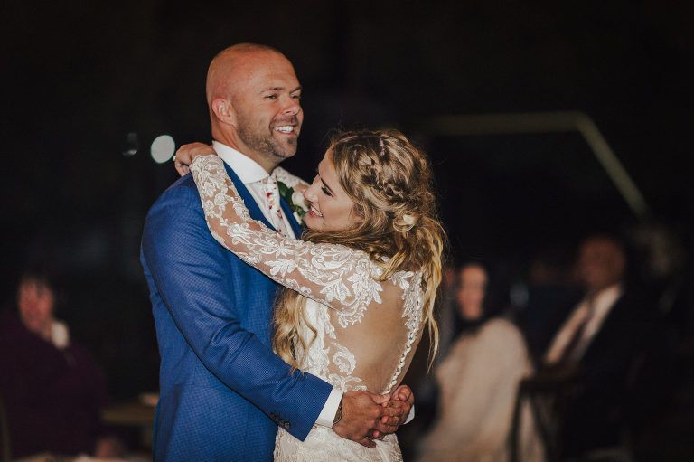 Couple dances at wedding reception; bride in lace dress, groom in blue suit, smiling.