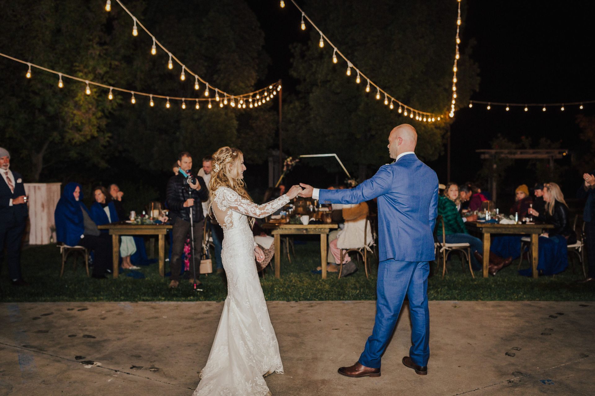 Bride and groom dancing under string lights at night, guests seated at tables in background.