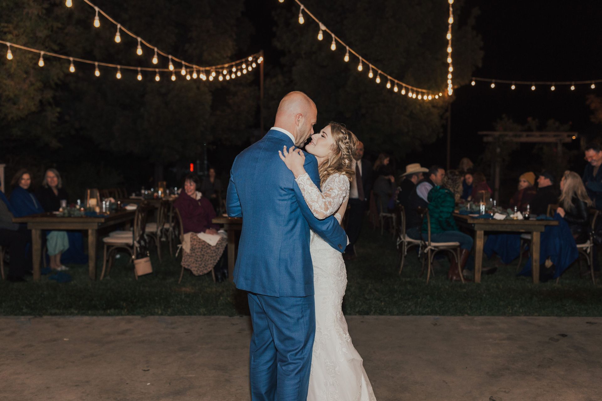 Bride and groom dance at night, lit by string lights. Guests watch at tables.