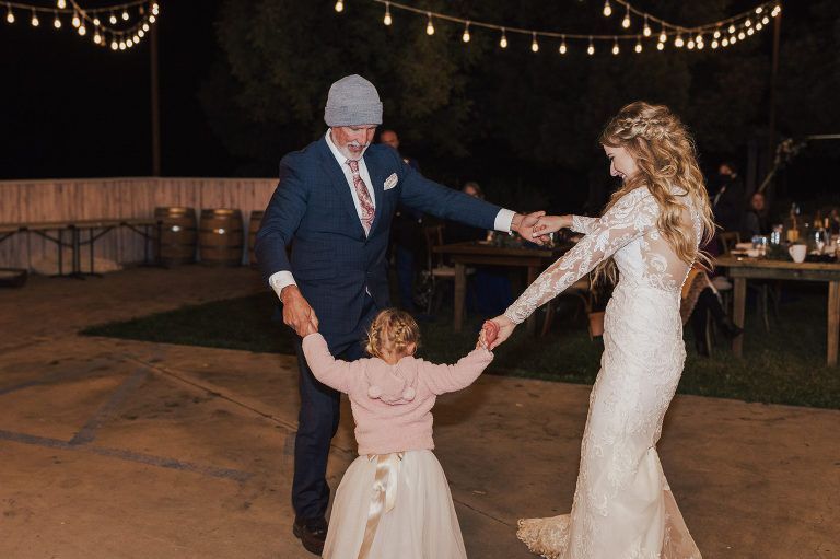 A man and bride dance with a child at an outdoor reception under string lights.