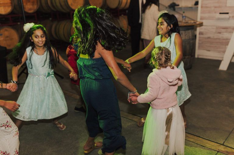 Children dancing in a circle, holding hands. They wear dresses and a jumpsuit, in a winery.