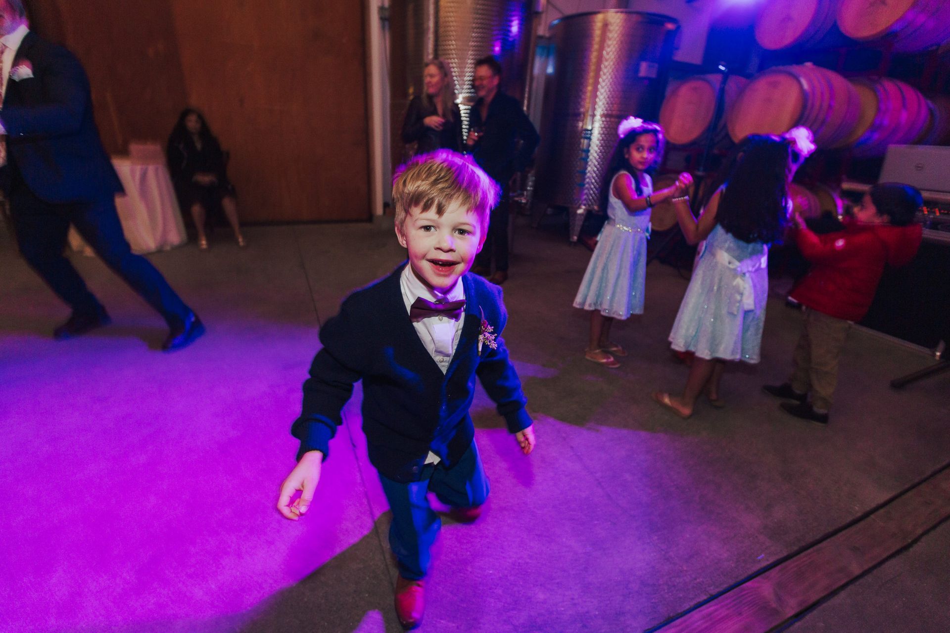 Boy in suit smiles, running toward camera at a party, other children dancing on purple-lit floor.