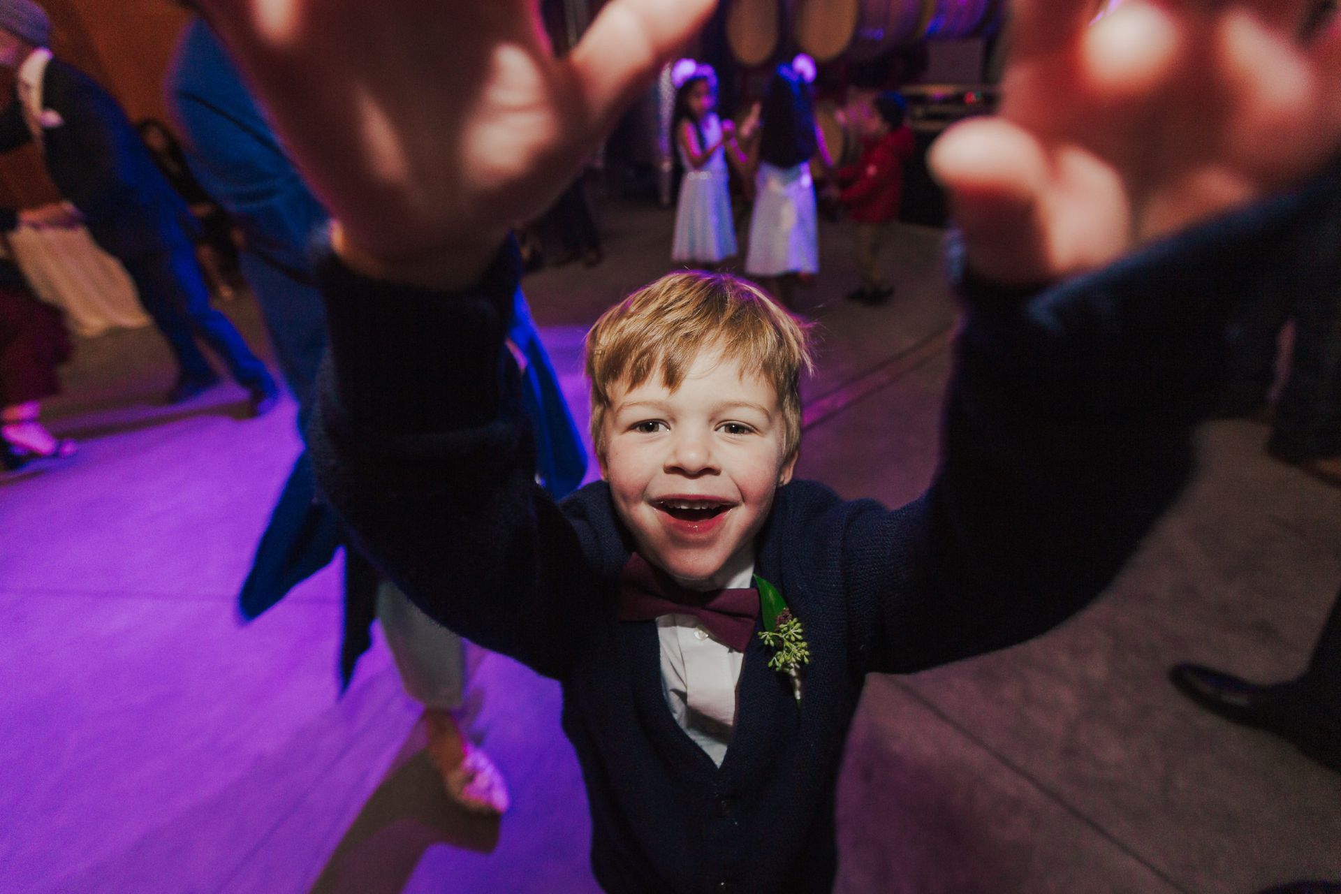 Boy in formal wear, arms raised, smiling widely on a dance floor with colorful lighting.