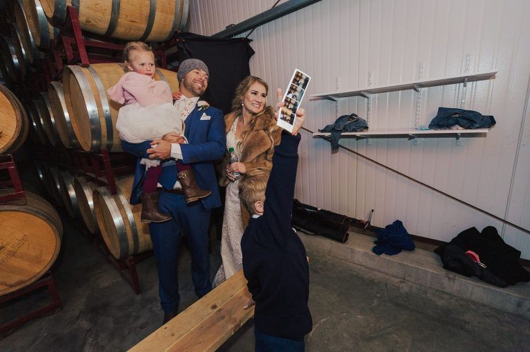 Bride and groom posing with child and photographer holding a photo strip, in a room with barrels.