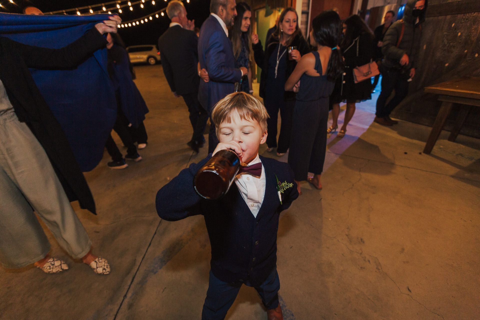 Boy in suit drinking from a large mug at an outdoor event with other guests.