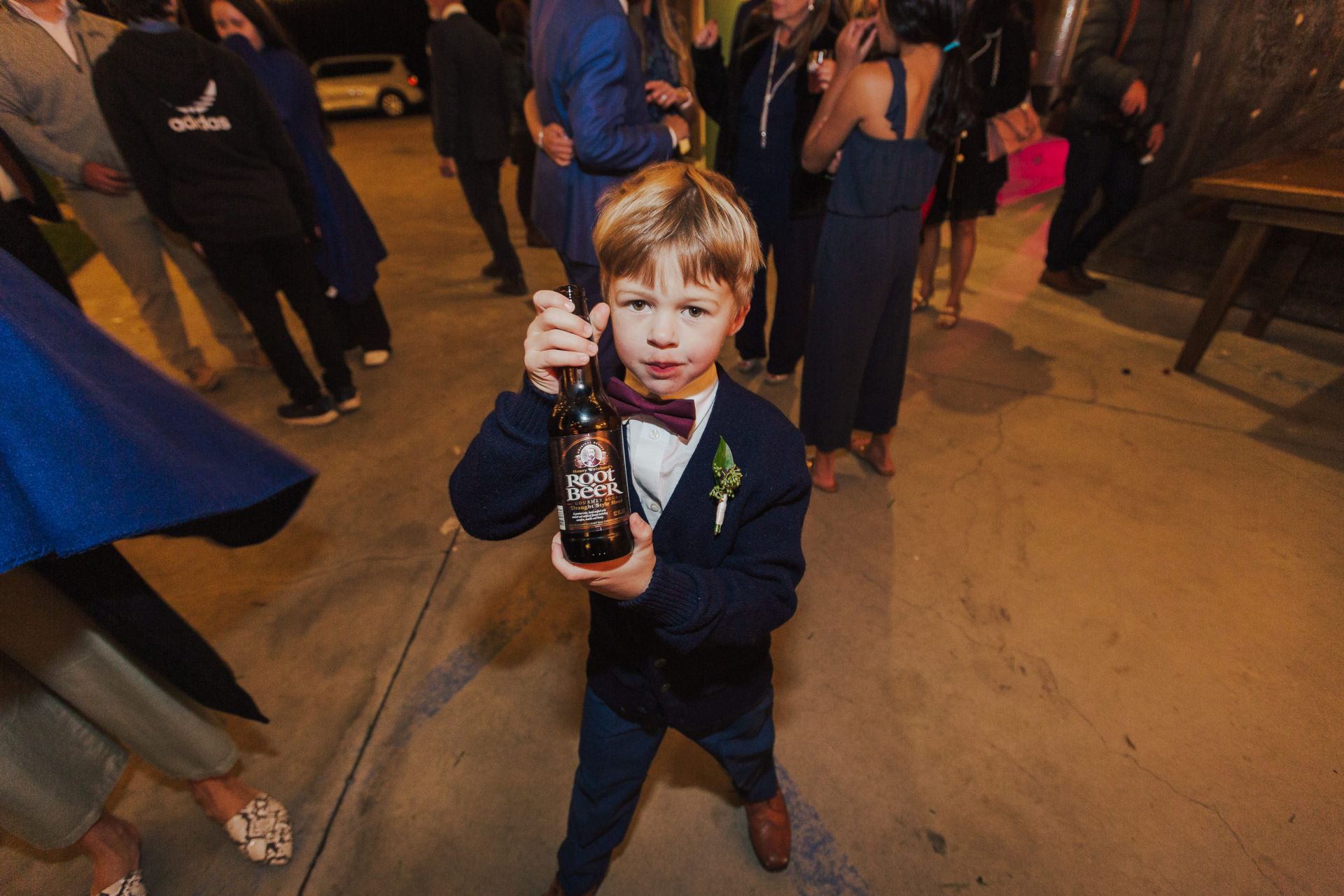 Boy in a suit holding a bottle, looking at the camera. He is at a party with others in the background.