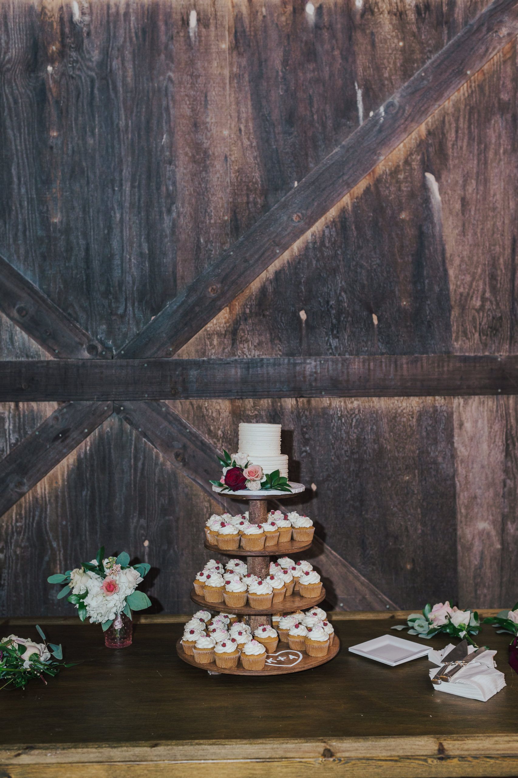 Cupcake tower with small wedding cake on top, on a wooden table. Barn door background.
