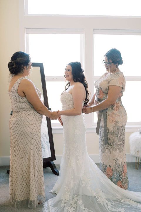 Bride in wedding dress, being helped by two women, in front of a window.