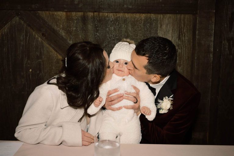 Parents kissing a baby wearing a white knit outfit and hat, seated at a table with a wooden background.