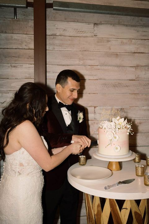 Couple cutting wedding cake on a small table. Bride in white dress, groom in burgundy tux. Light pink cake with flowers.