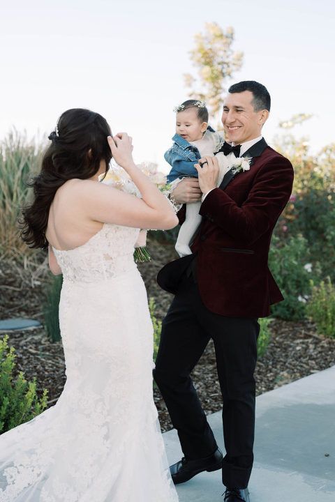 Bride adjusts hair, groom holds child; outdoor wedding celebration.