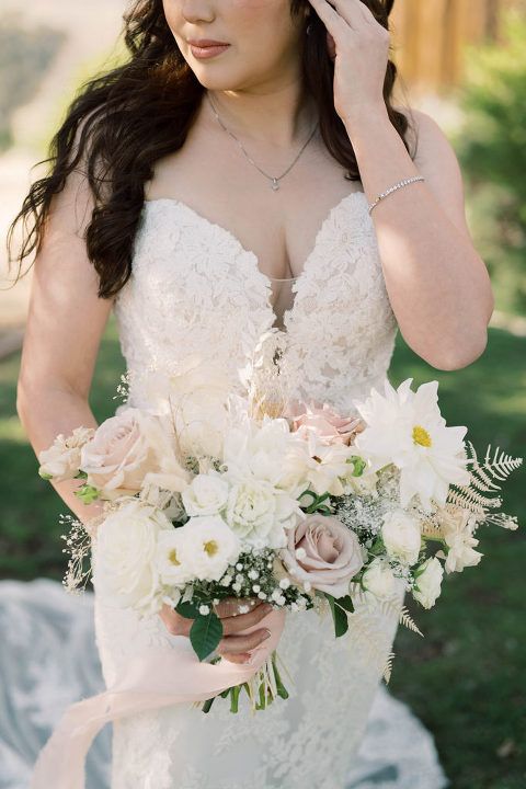 Woman in a white lace wedding dress holding a bouquet of white and blush flowers outdoors.