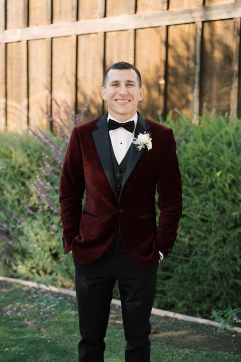 Man in a burgundy velvet tuxedo smiles, standing outdoors in front of a wooden fence and greenery.