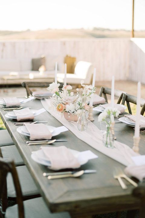 Wedding reception table set with flowers, candles, and blush napkins. Outdoor setting with sunlight.