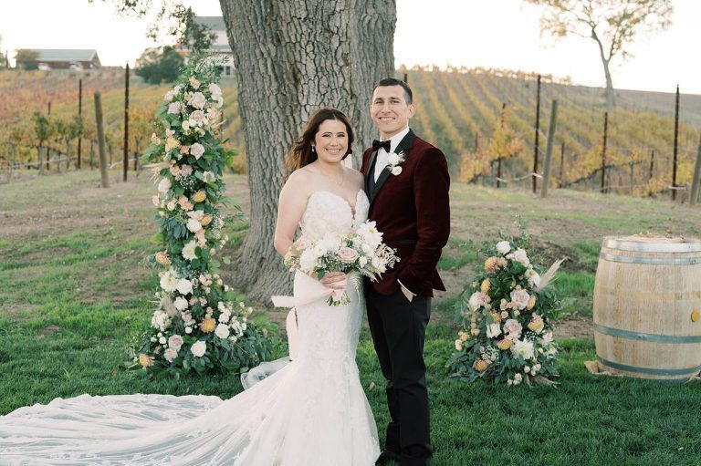Newlyweds posing in a vineyard, bride in white dress, groom in red velvet jacket, floral arch.