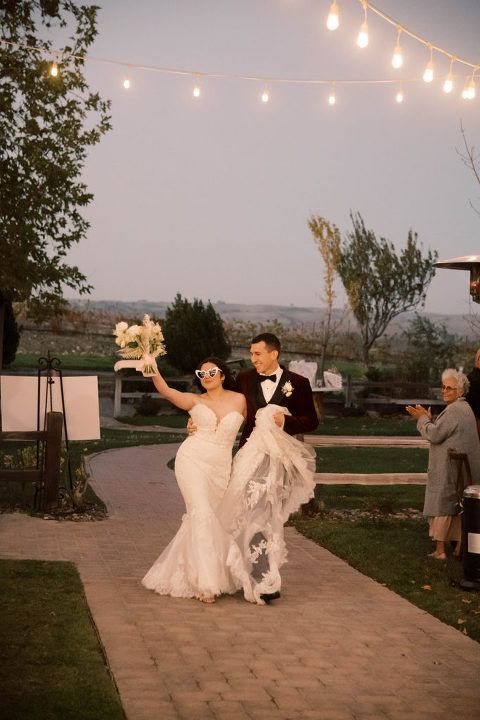 Newlyweds walking down a brick path, she waves her bouquet, he holds her train. Outdoor wedding with string lights.