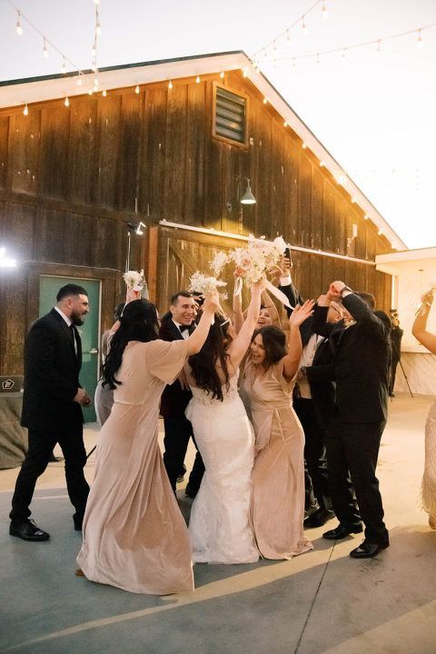 Wedding group cheering outdoors. Bride throws bouquet, bridesmaids and groomsmen celebrate near a wooden barn.