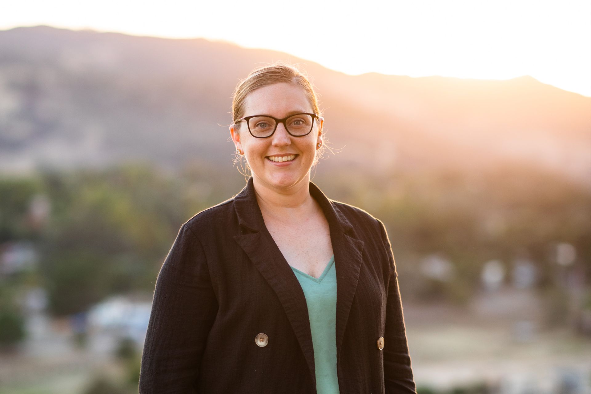Woman wearing glasses and blazer smiles, mountains in background, sunset light.