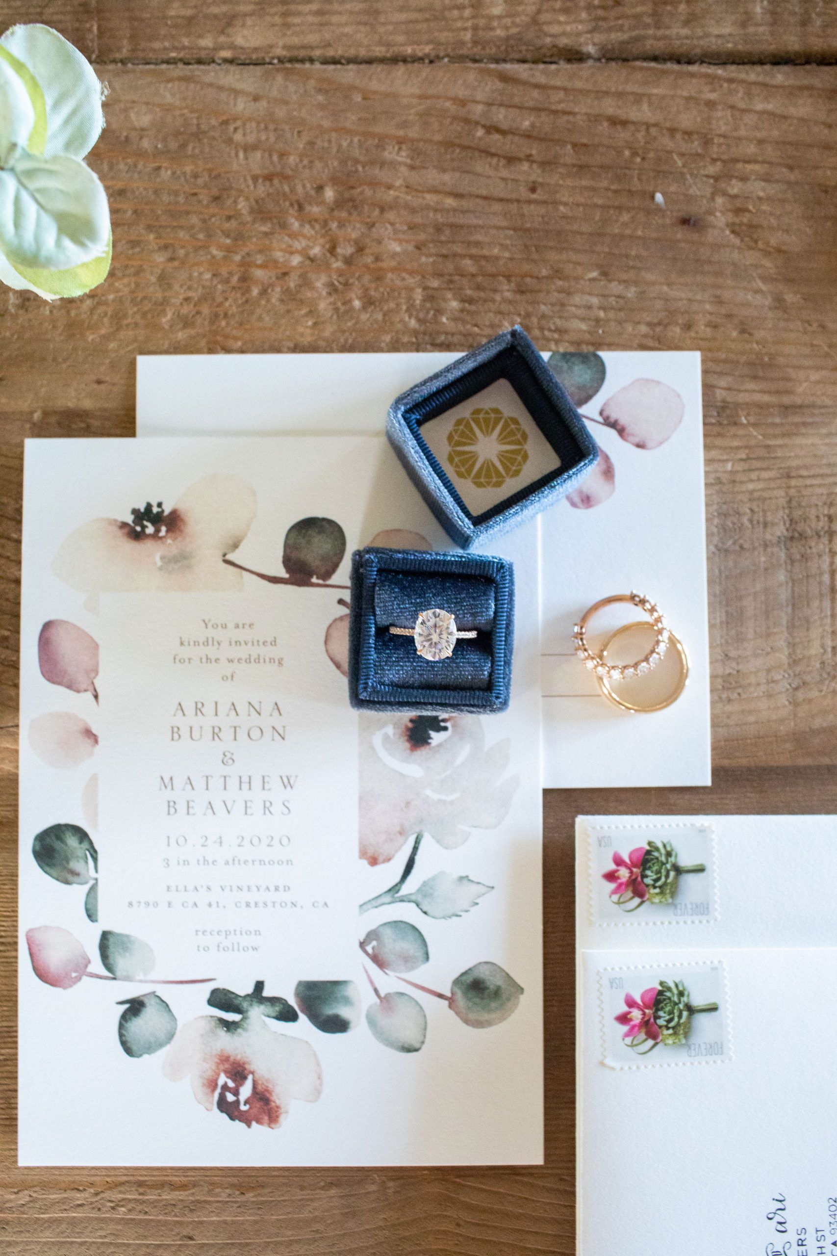 Wedding invitation with ring, and rings in velvet boxes on a wooden table with floral accents.