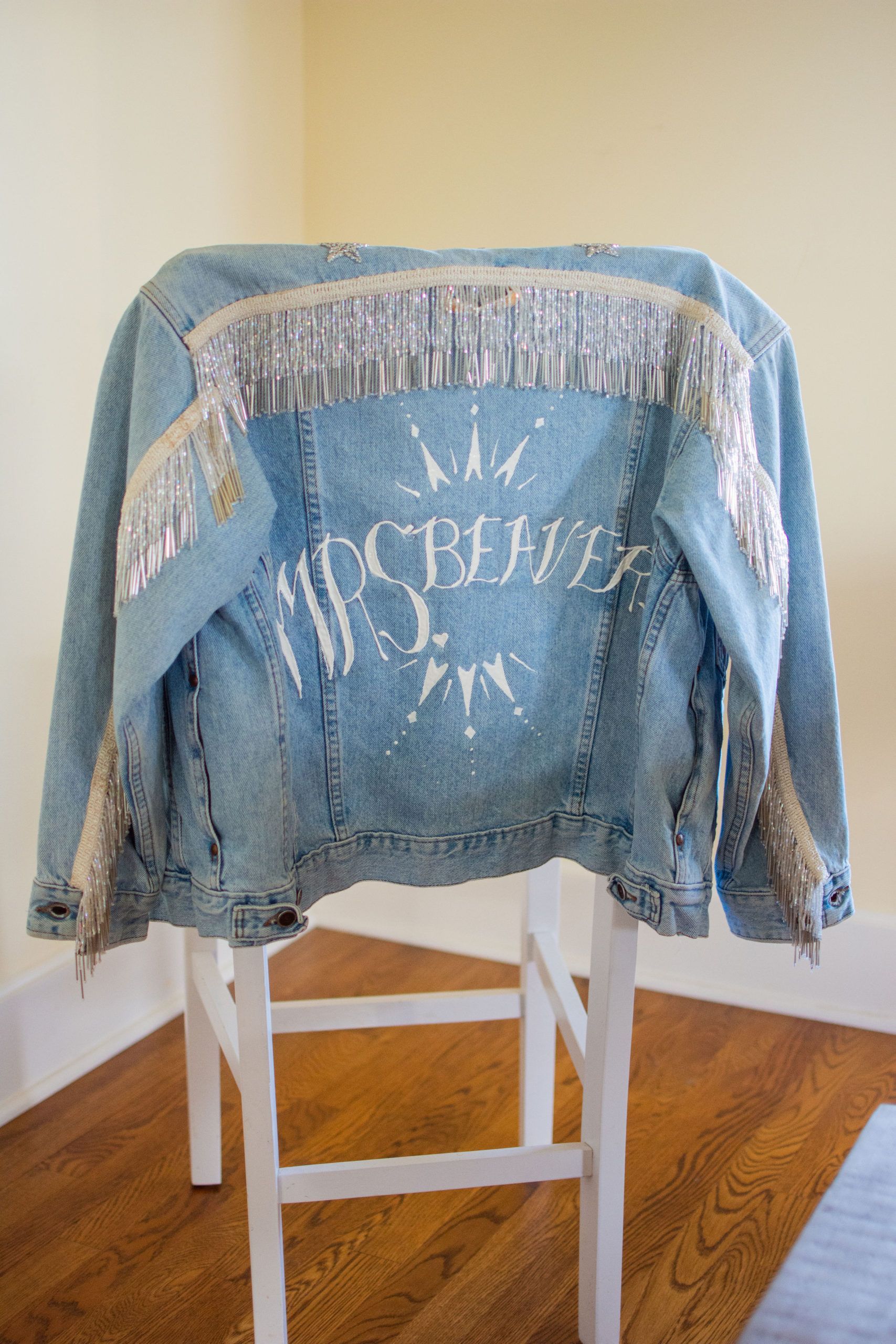 Light blue denim jacket with fringe and embellishments displayed on a white stool.